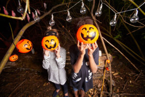 Two children holding plastic pumpkin buckets in front of their faces, with string lights hanging above them in a wooded area